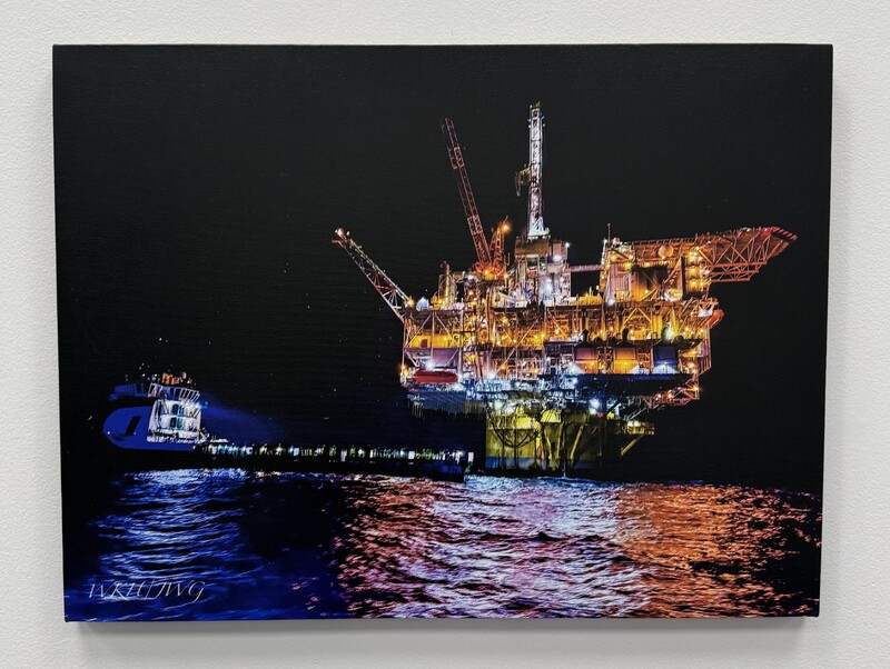 Offshore oil platform at night, illuminated with yellow and orange lights, with a spotlight from a nearby vessel reflecting on the ocean.