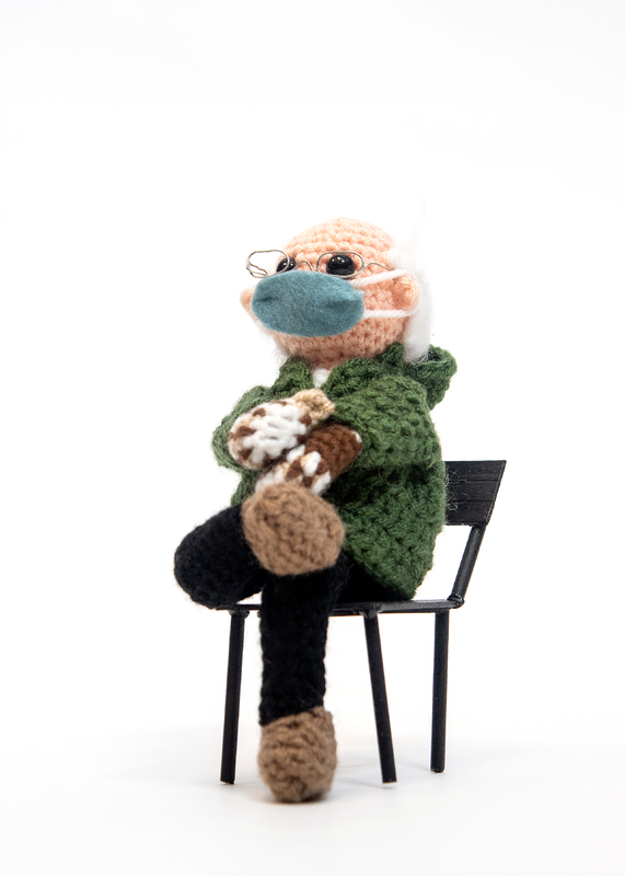 Crocheted man bundled in warm clothing including mittens seated in a chair.