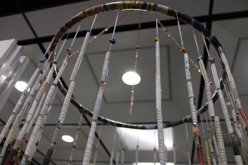 The installation consists of a circular frame from which numerous thin, colorful strips hang vertically. The strips appear to be made of tightly rolled paper, possibly from magazines or posters, and are suspended at various lengths, creating a curtain-like effect. T