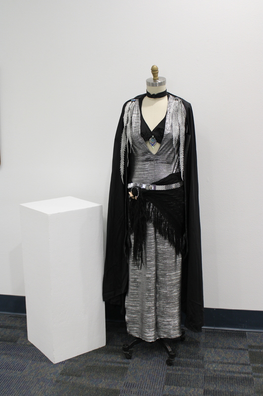 A 1930's Art Deco dance costume, consisting of a shiny silver gown, with a deep V-neck lined in black. A black cape with sheer, silver fern-like appliqués drapes over the shoulders. Around the waist, a silver sash decorated with sequins cinches the gown, accompanied by a black fringed overlay. A distinctive pendant hangs at the center of the chest, featuring a blue gem set within a geometric design.