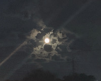 A full moon appears in a monochromatic night sky image with clouds. Two beams of moonlight or other reflections diagonally cross the image from bottom left to top right corner.  Shadowy images of power lines and trees can be seen towards the bottom of the image.