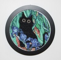 Embroidery hoop with black cat cross-stitch art in a green leafy background near blue irises.
