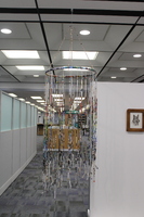 The installation consists of a circular frame from which numerous thin, colorful strips hang vertically. The strips appear to be made of tightly rolled paper, possibly from magazines or posters, and are suspended at various lengths, creating a curtain-like effect. 
