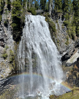 A waterfall cascades from a stony wall with trees and other vegetation. A rock bridge can be seen over the top of the falls. A rainbow is cast in an arc from left to right over the pool that the falls splash into.