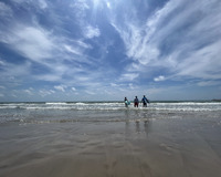 Three figures are standing on the beach in shallow surf. The figures are facing the water - away from the photographer. The two figures on the outside of the group carry surfboards. The blue sky has wispy white clouds and the bright sunlight is reflected in the waves.