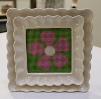 Cross-stitched pink flower with white center on a green background, framed in a wavy-edged cream ceramic frame.