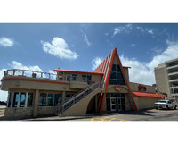 An outdoor picture of a building with a blue sky and bright white clouds in the background. The building is a two story structure built around a high peak roofline with vertical orange and white stripes. Yellow parking lot markings and a white pickup truck can be seen in front of the building.