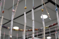 The installation consists of a circular frame from which numerous thin, colorful strips hang vertically. The strips appear to be made of tightly rolled paper, possibly from magazines or posters, and are suspended at various lengths, creating a curtain-like effect. T
