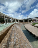 A large curved fountain, with water cascading down twelve steps into a channel surrounding a wide sidewalk around a raised grassy bed. The water comes from multiple fountain heads in a channel on top of the steps, that follow the curve. In the distance, stairs with handrails divide the fountain steps, which continue after the stairs. A covered canopy mirrors the curve beyond the fountain. Blue sky, white clouds and trees can be seen in the background of the image.