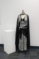  A 1930's Art Deco dance costume, consisting of a shiny silver gown, with a deep V-neck lined in black. A black cape with sheer, silver fern-like appliqués drapes over the shoulders. Around the waist, a silver sash decorated with sequins cinches the gown, accompanied by a black fringed overlay. A distinctive pendant hangs at the center of the chest, featuring a blue gem set within a geometric design.