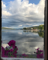 A calm body of water is viewed from a small window. Dark pink flowers are blooming under the window.  Buildings, trees, and mountains can be seen on the shorelines, visible to the right and left of the water. Mountains can be seen in the distance. Grey clouds with light tops hang low above the water and are reflected in its surface.