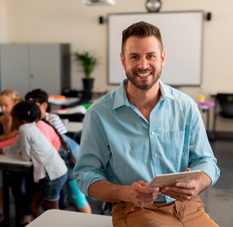 Image of a male teacher leaning against a desk with young students working behind.