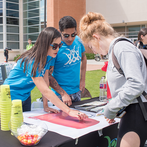 Two people in blue shirts outside at a table demonstrating a concept to a female student 